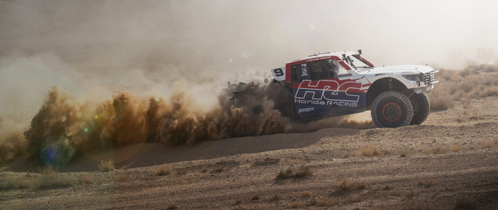 Red white and blue Desert race truck at full speed in the desert with a huge billowing plume of dust from the back wheels.